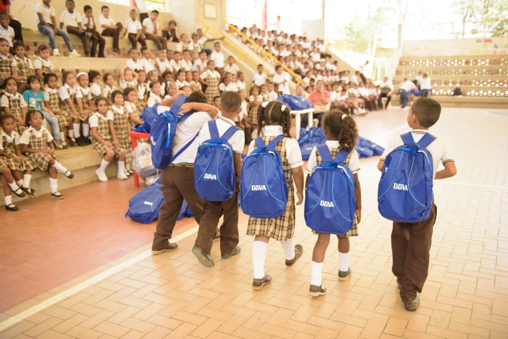 Niños caminan con morrales que tienen elementos escolares donados por BBVA Fotografía de niños caminando con morrales donados por BBVA con elementos escolares